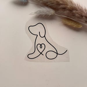 Könnte beinhalten: Ein schwarzer Line-Art-Aufkleber mit einer durchgehenden einzeiligen Zeichnung eines sitzenden Hundes, bei dem eine Herzform in den Brustbereich integriert ist. Das Design ist auf einer transparenten Klebefolie gedruckt und zeigt eine klare Silhouette.