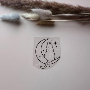 Könnte beinhalten: Ein schwarz-weißer Aufkleber mit einer Katzen-Silhouette, die auf einem Mond mit Sternen sitzt. Der Aufkleber befindet sich auf einer rosa Oberfläche, mit getrockneten Blumen im Hintergrund.