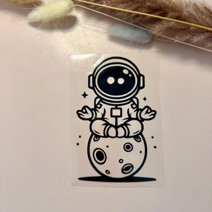 Puede incluir: Adhesivo en blanco y negro que representa a un astronauta en posición de meditación, sentado sobre una luna con cráteres. El astronauta lleva un traje espacial y un casco. Cuatro pequeñas estrellas rodean al astronauta y la luna.