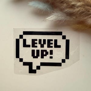 Könnte beinhalten: Ein schwarzer, pixeliger Sprechblasen-Aufkleber mit den Worten "LEVEL UP!" in fetten, weißen Buchstaben. Der Aufkleber hat einen transparenten Hintergrund, und das Design erinnert an Retro-Videospielgrafiken. Der Hintergrund ist hellrosa.