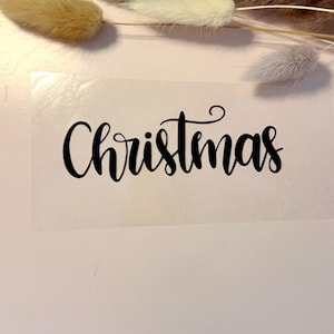 Könnte beinhalten: Ein transparenter Aufkleber mit dem Wort Christmas in einer schwarzen, fließenden Schreibschrift. Der Aufkleber ist unter mehreren getrockneten, flauschigen, beigen und hellgrauen Ziergrasstängeln positioniert.