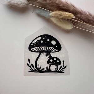 Peut inclure: Autocollant de champignon noir et blanc représentant deux champignons avec des chapeaux à pois et des tiges détaillées. L'autocollant a une bordure transparente et est entouré de petits détails en forme de feuilles. Le design est une illustration fantaisiste.