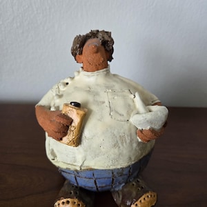 Puede incluir: Una figurita de cerámica de una persona que lleva una camisa blanca con pantalones azules a cuadros y sostiene una bolsa de papel marrón y un tazón blanco.