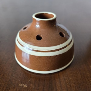 May include: A small, brown ceramic flower frog with white stripes. It has five holes in the top for holding flowers.