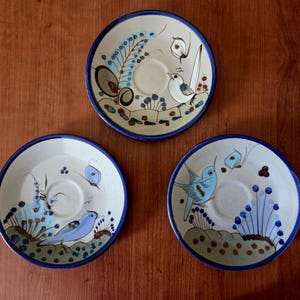 May include: Three round ceramic saucers with a cream-colored base and a dark blue rim. Each saucer features hand-painted designs of blue birds, butterflies, and floral elements in shades of blue, brown, and green, arranged around a central indentation.