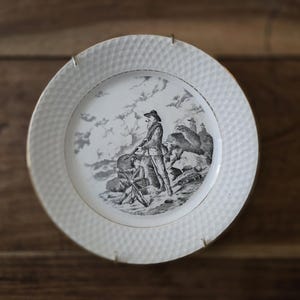 May include: White porcelain plate with a diamond pattern around the rim and a black and white illustration of a man and a boy on a rocky landscape. The plate has a gold trim.