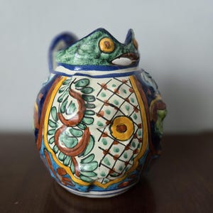 Puede incluir: Jarra de cerámica colorida con diseño de pez. La jarra tiene una parte superior verde y azul, un asa y está adornada con intrincados patrones en tonos azules, amarillos, rojos y blancos. La jarra es un objeto decorativo.
