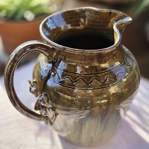 May include: A handcrafted ceramic pitcher with a dark brown glaze and a decorative wavy pattern around the top. The pitcher has a curved handle and a spout for pouring. The base of the pitcher has a light green and brown gradient.