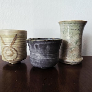 May include: Three handcrafted ceramic vessels in earthy tones. The leftmost cup is beige with brown markings. The center cup is dark grey. The rightmost vase is light green and beige. All are on a dark brown surface.
