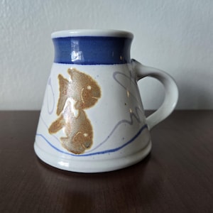 May include: White ceramic mug with a blue rim and a gold fish design. The mug has a handle and is sitting on a wooden surface.