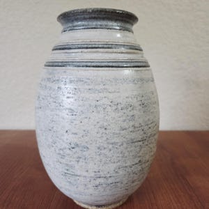 May include: A handmade ceramic vase with a textured, speckled white and grey finish. The vase features horizontal bands of grey and a dark grey rim. The vase is a decorative item for the home.