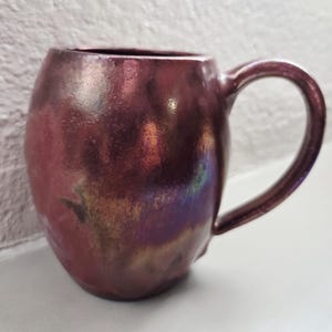 Może przedstawiać: Ręcznie wykonany, iryzujący ceramiczny kubek o zaokrąglonym korpusie i dużym uchu. Kubek ma głęboki bordowy kolor z nutami złota i zieleni, tworząc efekt połysku. Kubek jest przeznaczony do gorących napojów.