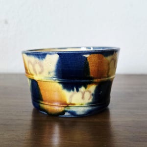 Pode incluir: Uma pequena tigela de cerâmica artesanal com um design único. A tigela apresenta um exterior azul escuro com manchas de amarelo e castanho. O interior é de um tom de azul mais claro. A tigela tem uma forma cilíndrica com uma borda ligeiramente alargada.
