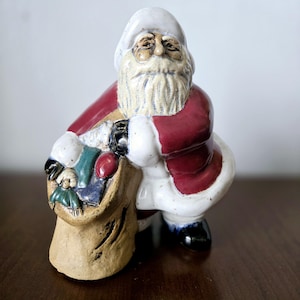 Puede incluir: Una figura de cerámica de Papá Noel con barba y gorro blancos, lleva un abrigo rojo y sostiene un saco marrón lleno de regalos coloridos.