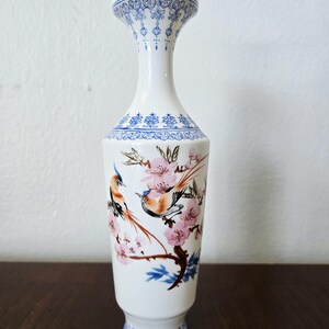 Può includere: Vaso in ceramica bianca con collo sottile e base svasata. Il vaso presenta un motivo floreale blu nella parte superiore e inferiore, con un disegno centrale di uccelli appollaiati su rami fioriti con fiori rosa. Un pezzo decorativo per la casa.