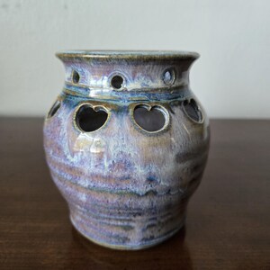 Może przedstawiać: Mały, ręcznie robiony ceramiczny garnek z szkliwioną powierzchnią w odcieniach fioletu, błękitu i brązu. Garnek ma okrągły korpus z szerokim otworem i szereg sercowatych wycięć wokół środka.