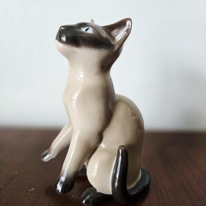 May include: A ceramic Siamese cat figurine in a seated pose, looking upwards. The cat has a cream-colored body with dark brown markings on its face, ears, paws, and tail. The figurine has blue eyes and is on a dark brown surface.