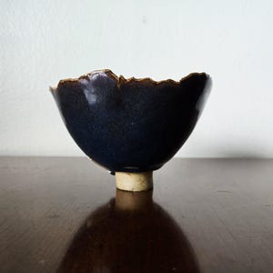 May include: A small, dark blue ceramic bowl with a rough, textured rim. The bowl has a flared shape and sits on a short, cylindrical, beige base. The interior of the bowl is a deep, glossy blue.