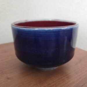May include: A small, round ceramic bowl featuring a deep, glossy navy blue exterior glaze that transitions to a lighter blue near the rim. The interior of the bowl is finished with a smooth, solid dark red glaze, creating a contrast.