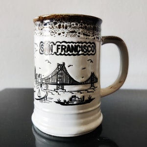 Może przedstawiać: Biały kubek ceramiczny z brązową obwódką i czarno-białym rysunkiem mostu Golden Gate oraz napisem "SAN FRANCISCO".