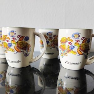 Könnte beinhalten: Vier weiße Keramikbecher mit grünen Rändern. Jeder Becher zeigt eine farbenfrohe Illustration von Meeresschildkröten und Fischen. Die Becher sind mit dem Text "Coronado, Kalifornien" bedruckt.