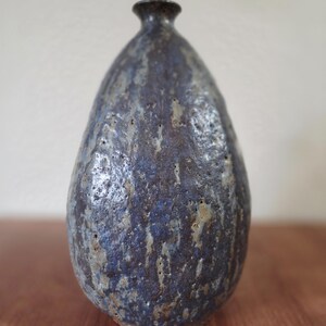 Puede incluir: Un jarrón de cerámica hecho a mano con un esmalte moteado azul oscuro y marrón. El jarrón tiene un cuerpo redondeado y un cuello estrecho, de unos 25 cm de altura. La superficie tiene un aspecto texturizado y orgánico.