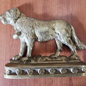 May include: A bronze-colored dog figurine in a standing pose. The dog has a bell around its neck and is mounted on a rectangular base with a decorative border. The sculpture is detailed and appears to be made of metal.