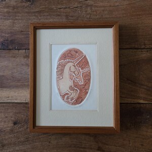 Puede incluir: Obra de arte enmarcada con un relieve de un unicornio. El unicornio se representa de perfil, con una melena fluida y un cuerno en espiral. La obra de arte está sobre un fondo ovalado con una apariencia texturizada, de color marrón rojizo, enmarcada en madera.