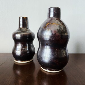 Peut inclure: Deux vases en céramique brun foncé avec une finition brillante. Les vases ont une forme bulbeuse avec un col étroit et une base blanche. Le plus grand vase mesure environ 20 cm de haut, et le plus petit vase mesure environ 15 cm de haut.