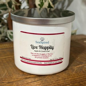 Op de afbeelding: Een witte kaars met een zilveren deksel, met de tekst "Live Happily" en "Apple & Green Leaf". Het etiket bevat het merk "SeaSpired" en het nettogewicht van 482g. De kaars staat op een houten oppervlak.