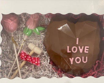 May include: A chocolate heart-shaped box with the words "I love you" in pink lettering. The box is surrounded by pink and white tissue paper and includes two pink lollipop roses and a small wooden hammer.