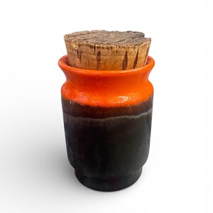 May include: A small, ceramic jar with a cork stopper. The jar is cylindrical with a flared top, featuring a gradient of colours: orange at the top, transitioning to black at the base. The cork stopper is a rough, natural wood.
