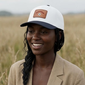 Puede incluir: Gorra de béisbol blanca con visera azul marino y parche de cuero marrón. El parche presenta la palabra "Grenadiye" sobre un gráfico de balón de fútbol. La gorra la lleva una persona con una blazer color beige.