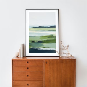 May include: Abstract landscape painting in shades of green and blue, framed in black, hanging above a wooden dresser with drawers.