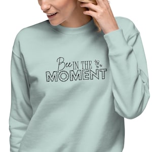 May include: A light sage green long-sleeve sweatshirt featuring the black text "Bee IN THE MOMENT" printed across the chest. The word "Bee" is in a cursive script, while "IN THE MOMENT" is in a bold, blocky font with a honeycomb pattern inside.