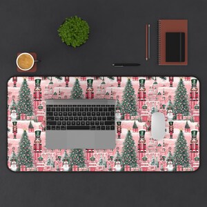 May include: A pink and white desk mat with a repeating pattern of nutcracker soldiers, Christmas trees, and snowmen. A laptop computer and a computer mouse are on the mat.