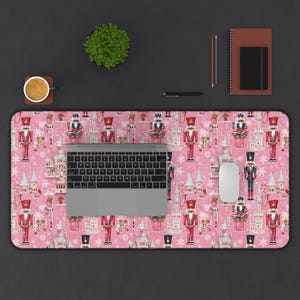 May include: Pink desk mat with a repeating pattern of nutcracker soldiers, snowflakes, and candy canes. A laptop computer and a mouse are on the mat.