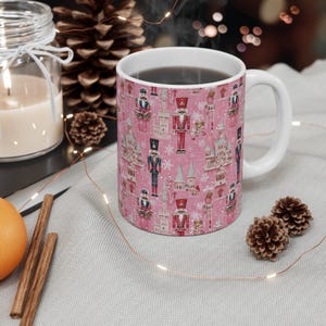 May include: A pink ceramic mug with a Christmas nutcracker pattern. The mug is filled with a dark liquid and has a white handle.