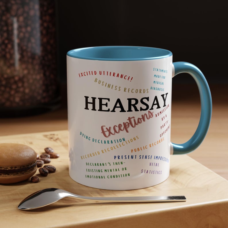 May include: White ceramic coffee mug with a blue rim and handle. The mug features a graphic design with the word "HEARSAY" in large letters and the word "Exceptions" in smaller letters. The graphic also includes a list of legal terms related to hearsay exceptions.