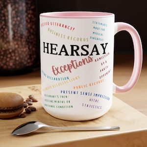 May include: White ceramic mug with a pink rim and handle. The mug features the word "HEARSAY" in black text and the word "Exceptions" in pink text. The mug also features a list of legal terms related to hearsay exceptions, including "Excited Utterance", "Business Records", "Statements Made for Medical Diagnosis", "Dying Declaration", "Recorded Recollections", "Admission by a Party-Opponent", "Public Records", "Present Sense Impression", "Declarant's Then-Existing Mental or Emotional Condition", and "Vital Statistics".