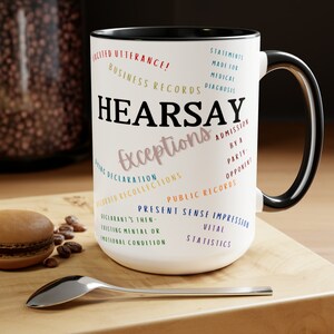 May include: White ceramic mug with a black handle. The mug has the word "HEARSAY" printed in black with the word "Exceptions" printed in pink below it. The mug is surrounded by text in various colors, including "Business Records", "Statements Made for Medical Diagnosis", "Dying Declaration", "Recorded Recollections", "Admission by a Party", "Opponent", "Public Records", "Present Sense Impression", "Declarant's Then-Existing Mental or Emotional Condition", and "Vital Statistics".