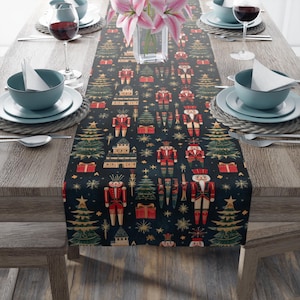 May include: A black table runner featuring a repeating pattern of nutcrackers, Christmas trees and stars. The nutcrackers are wearing red and gold uniforms and holding a weapon. The Christmas trees are green and gold. The stars are gold.