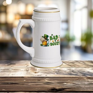 以下が含まれることがあります： 緑のクローバーのデザインと「Bad and Boozy」の文字が書かれた白い陶器製のビアジョッキです。