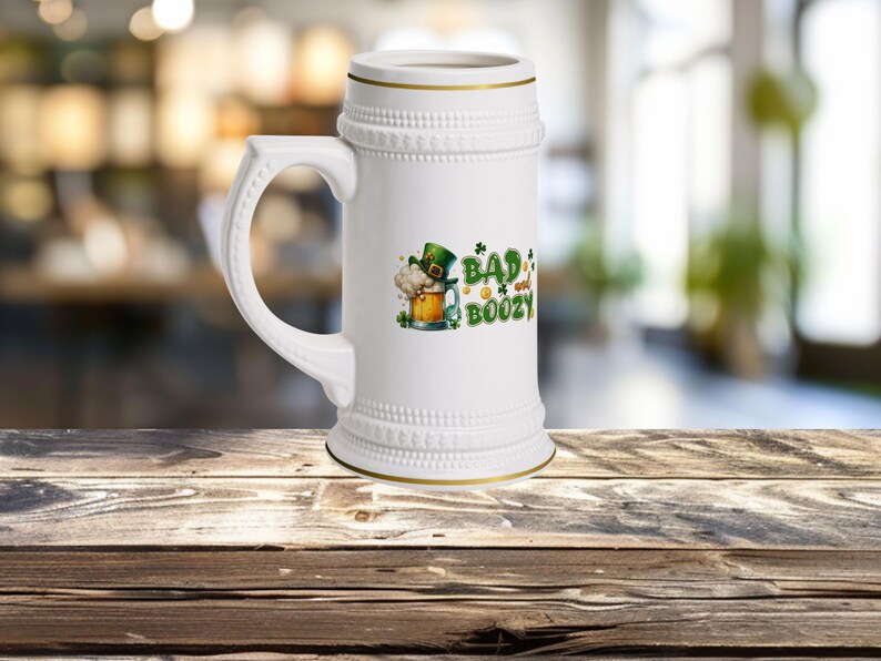 以下が含まれることがあります： 緑のクローバーのデザインと「Bad and Boozy」の文字が書かれた白い陶器製のビアジョッキです。