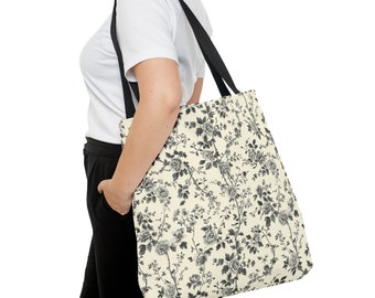 May include: A cream-colored tote bag with a black floral pattern. The bag has black straps and is being held by a person.