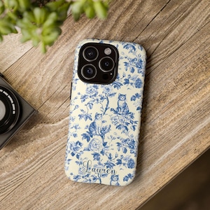 Peut inclure: Coque de téléphone avec un fond crème et un motif floral et chat bleu. Le motif répète des fleurs, des feuilles et des chats. Le nom "Lauren" est écrit en cursive en bas de la coque.