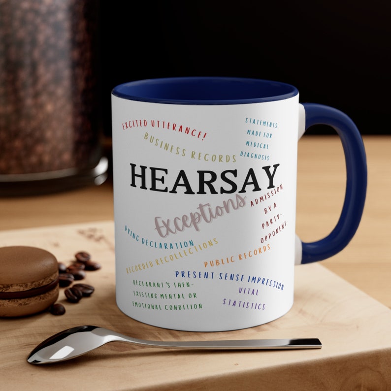May include: White ceramic coffee mug with a blue rim. The mug features the word "HEARSAY" in large, colorful letters, with the word "Exceptions" written in a smaller font below. The mug is decorated with a variety of legal terms related to hearsay exceptions, including "Dying Declaration", "Recorded Recollections", "Public Records", and "Present Sense Impression".