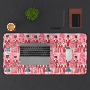 May include: Pink and white desk mat with a repeating pattern of nutcracker figurines holding presents and snowflakes. A laptop computer and a white mouse are on the mat.