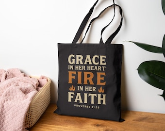 May include: Black tote bag with white text that reads "Grace in her heart Fire in her Faith Proverbs 31:26". The text is in a vintage style font with orange flames on either side of the word "Fire".