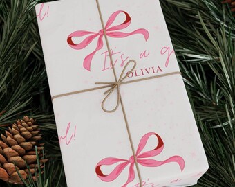 May include: White gift wrap with pink bows and the text "It's a girl! Olivia" printed on it. The wrap is tied with twine.